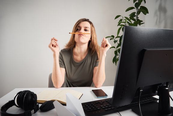Eine junge Frau sitzt an einem Tisch mit einem Laptop und einem Notizbuch. Sie trägt Kopfhörer und hält einen Stift zwischen den Lippen, während sie konzentriert ist.